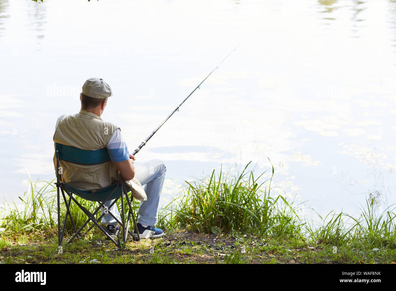 rear-view-of-mature-fisherman-sitting-on-chair-with-fishing-rod-and-waiting-for-a-catch-at-the...jpg